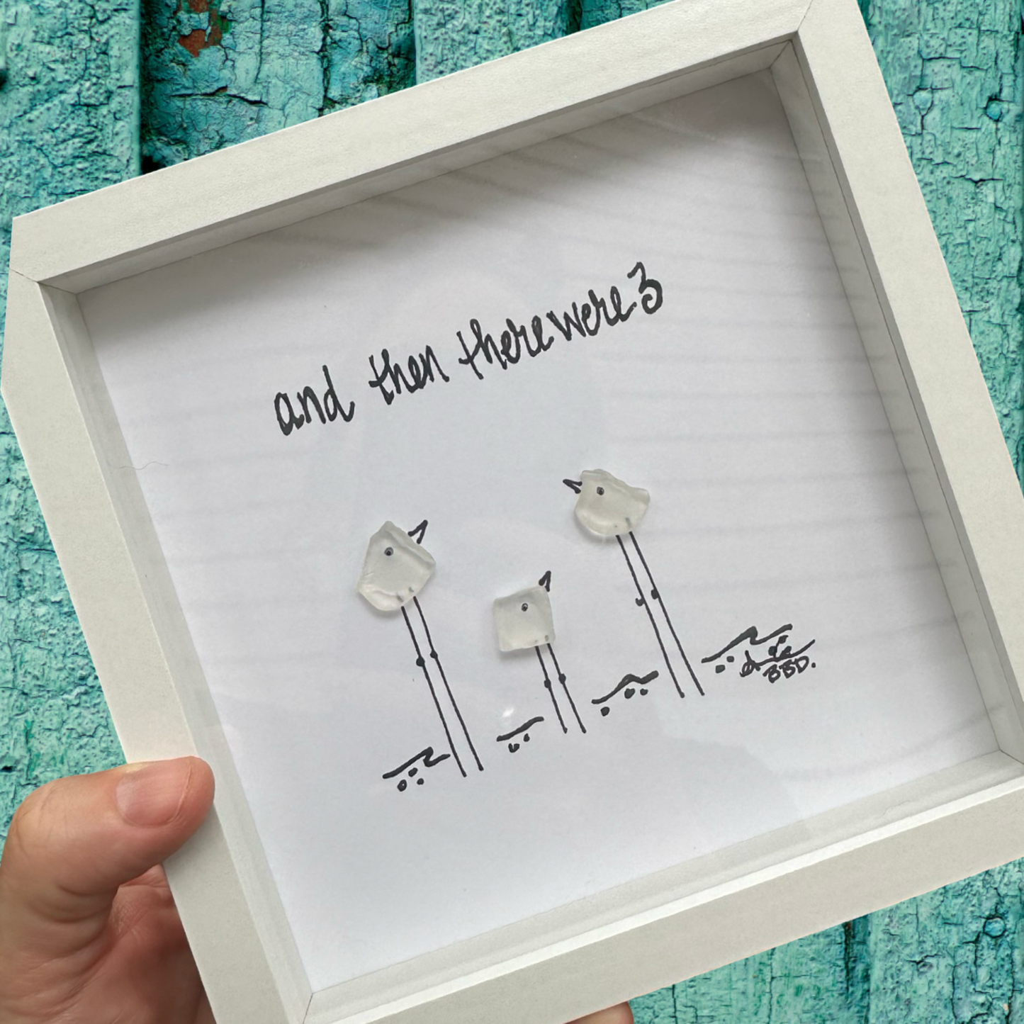 A framed artwork depicting three birds perched on a branch, with the text 'And then there were 3' at the top, held in a person's hand against a blue wooden background.