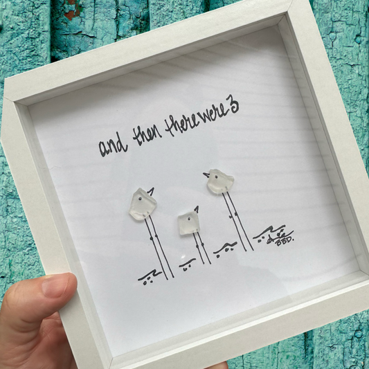 A framed artwork depicting three birds perched on a branch, with the text 'And then there were 3' at the top, held in a person's hand against a blue wooden background.