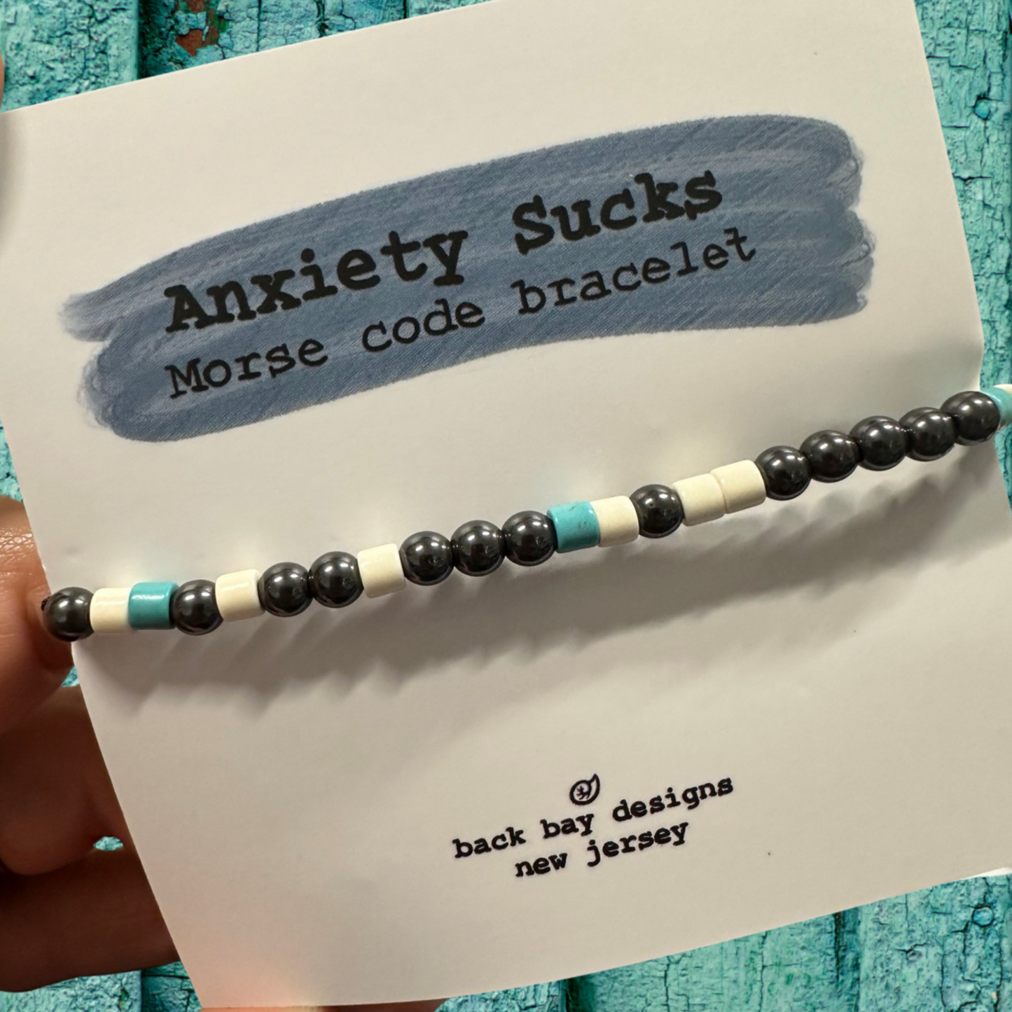 Beaded bracelet labeled 'Anxiety Sucks Morse code bracelet' on a card with text.