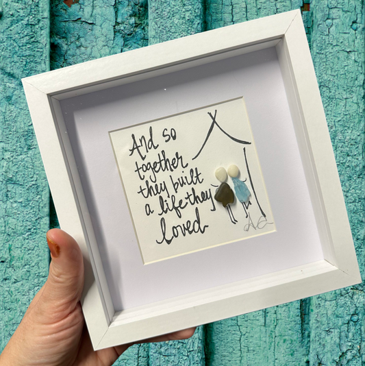 A 6x6 shadowbox with plexiglass, featuring a written message 'And so they built a life together' in a black script, with a small decorative figure inside.