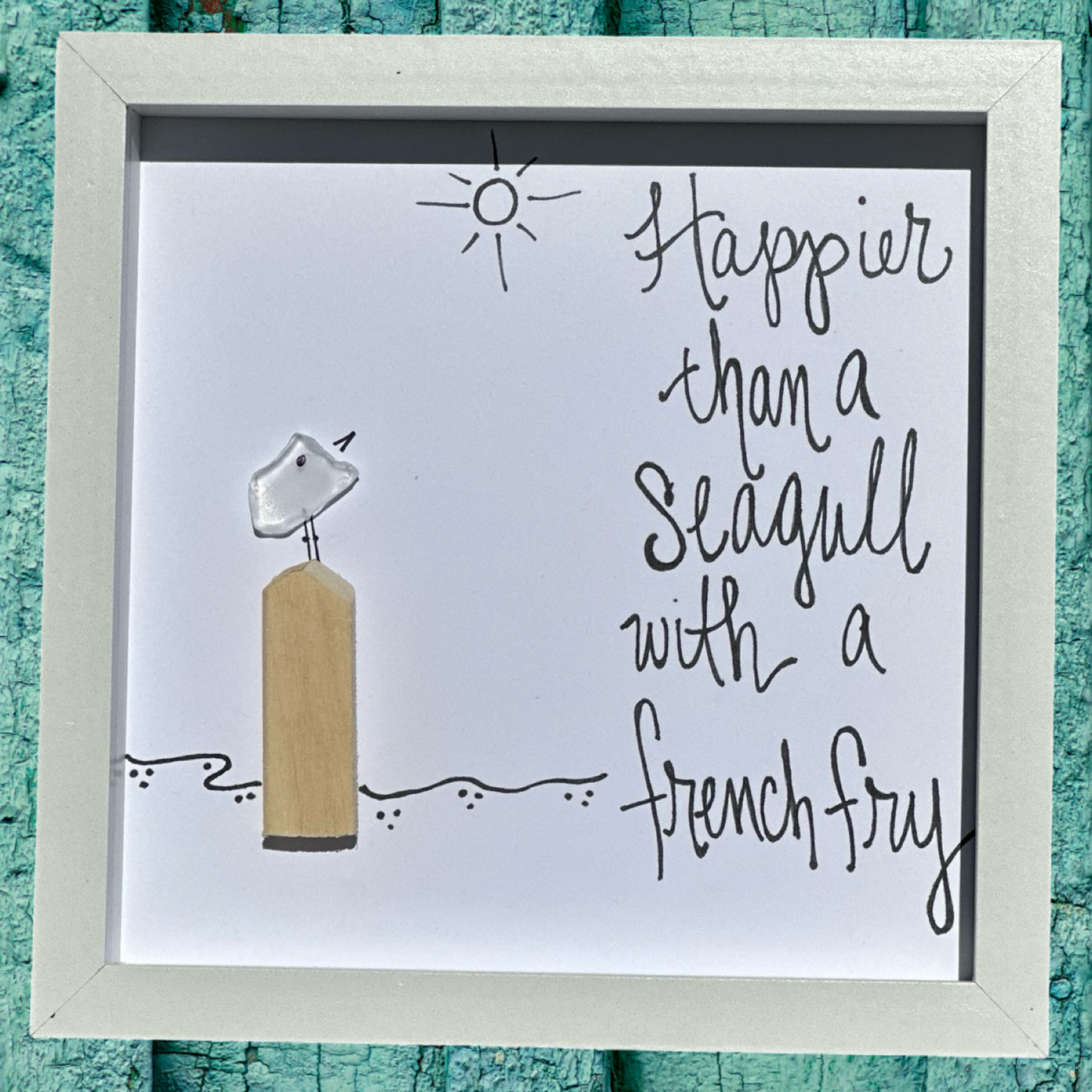 A framed shadowbox art piece with a seagull and a piece of seaglass, surrounded by a sun and ocean background, with the text 'Happier than a seagull with a French fry'.