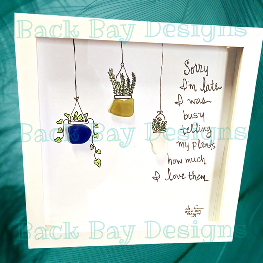 An 8 x 8 shadowbox frame with seaglass art, featuring a variety of seaglass plants and flowers, and the text 'Sorry I'm late I was busy telling my plants how much I love them'. sea glass art by back bay designs