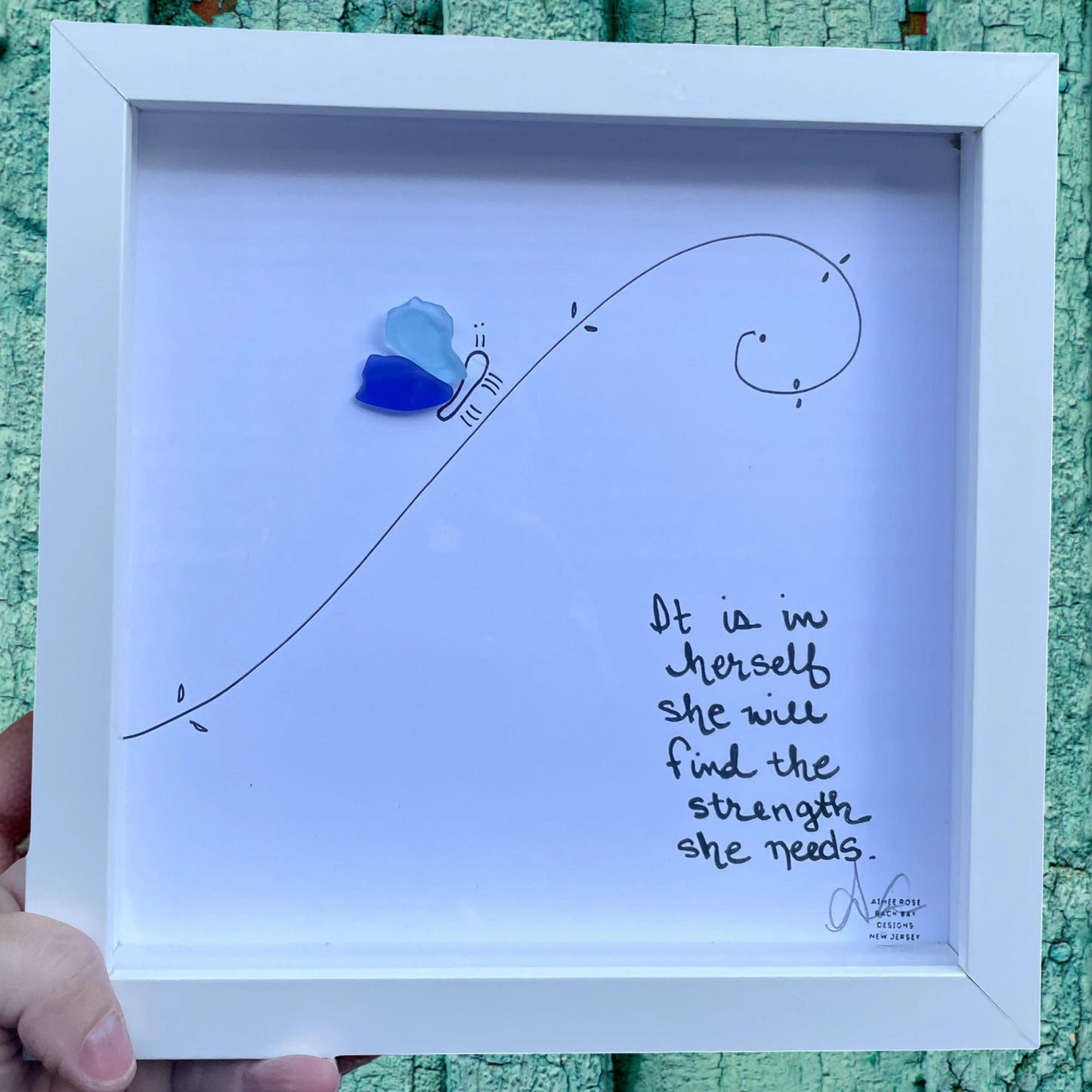 Framed artwork with a blue butterfly and handwritten text on a white background, held against a textured green surface. It is in herself she will find the strength she needs.