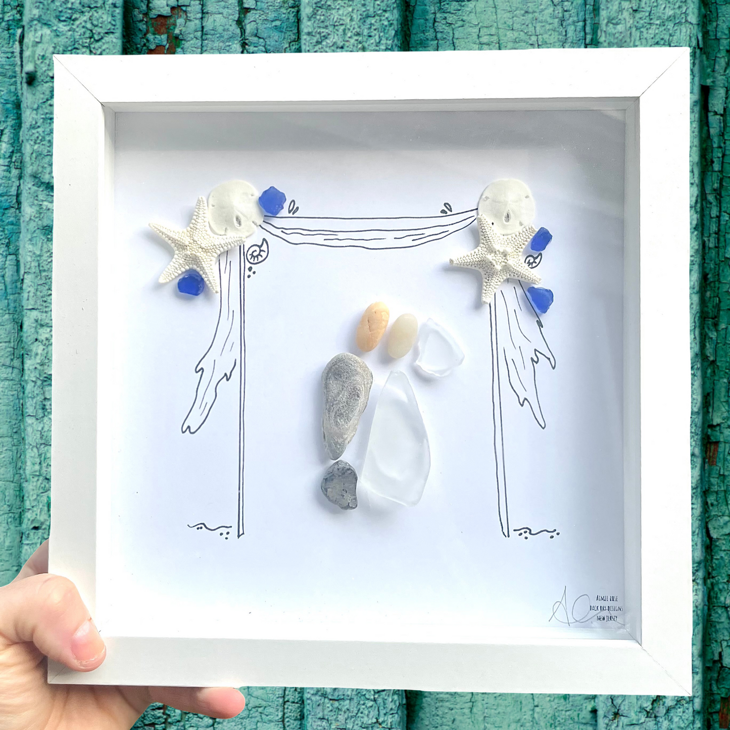 Framed wedding artwork with starfish, shells, and pebbles on a white background against a teal wooden surface.