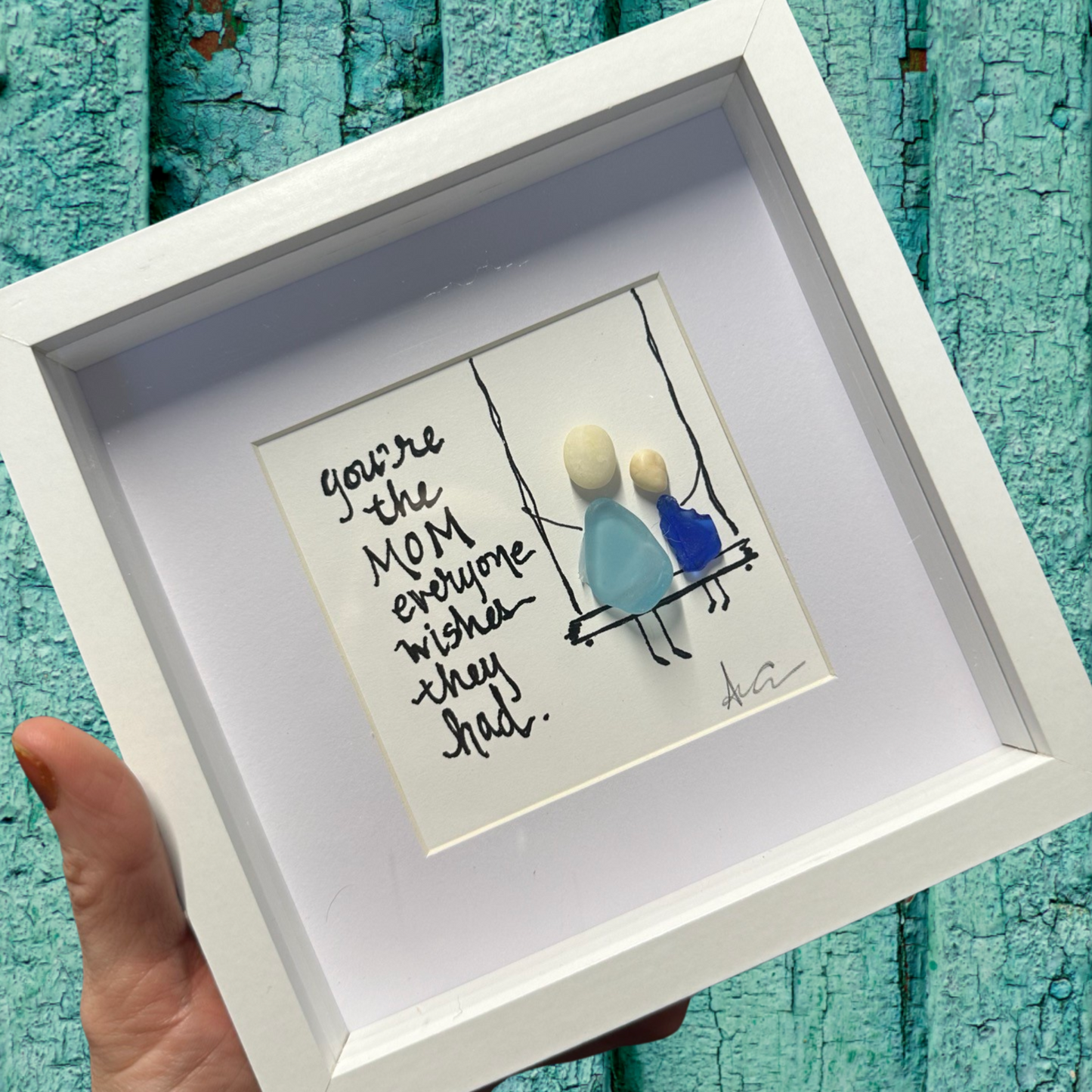 A shadow box frame with plexiglass, holding a seaglass art piece with a written message that reads 'You're the mom everyone wishes they had'. The frame is white and has a visible signature at the bottom.