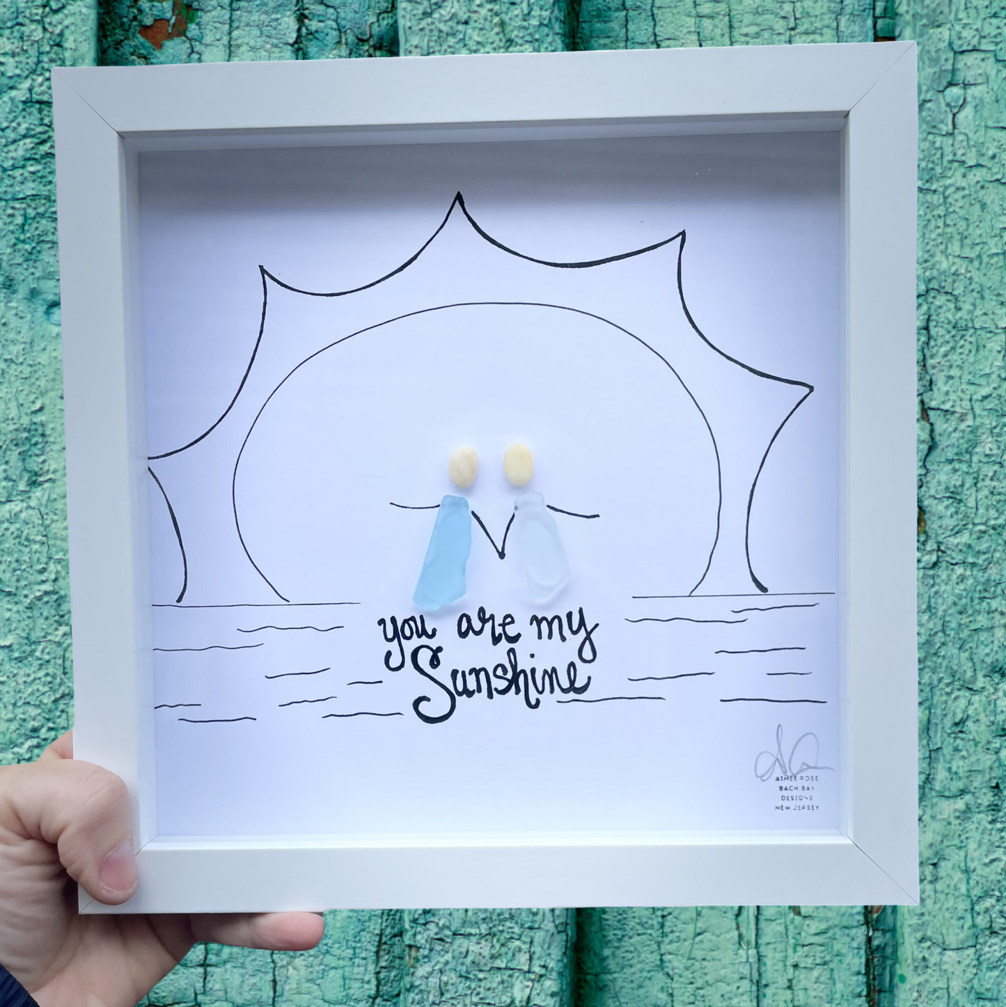 Framed drawing of a sun and a child with 'you are my sunshine' text against a textured teal background.