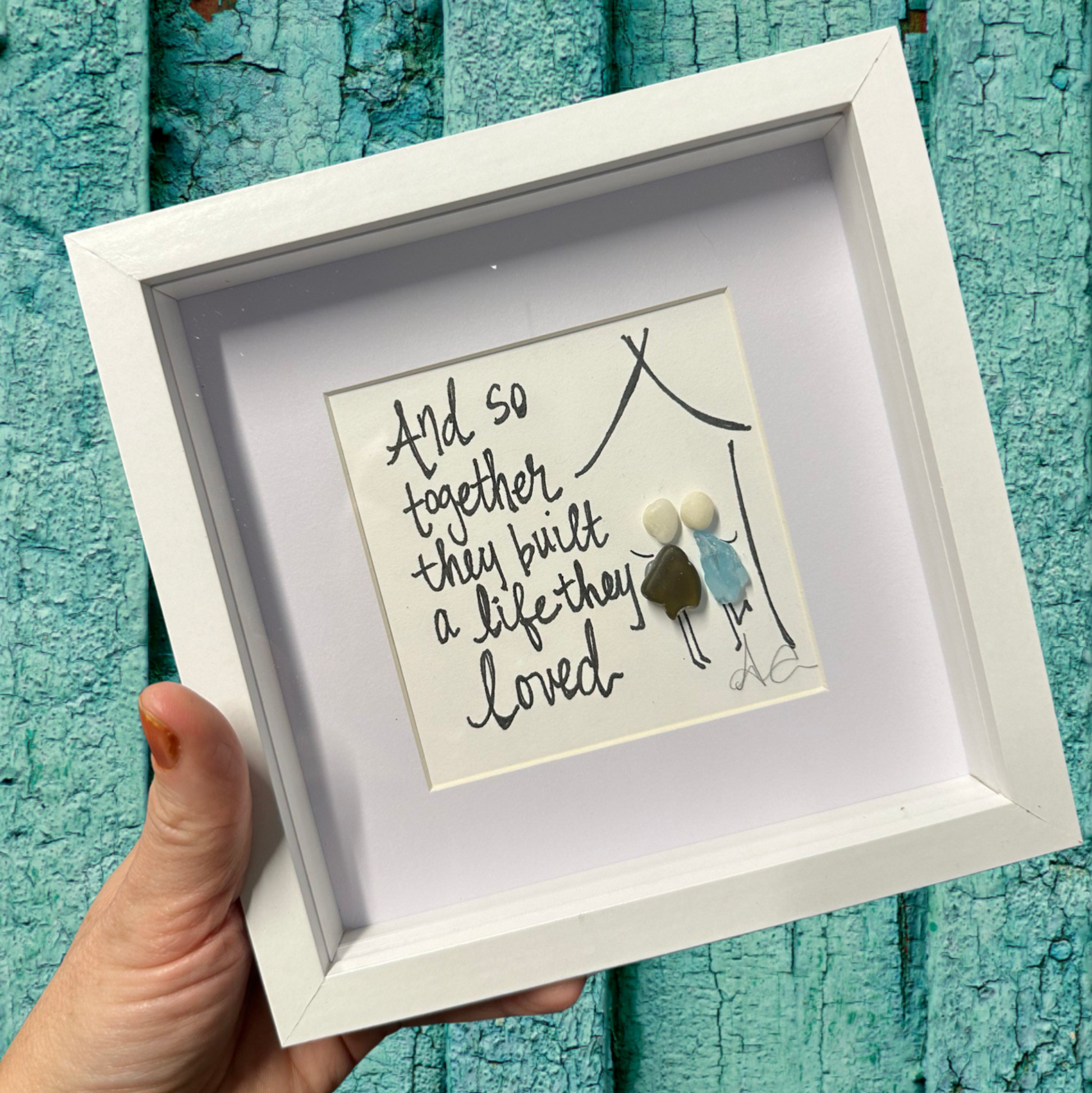 A 6x6 shadowbox with plexiglass, featuring a written message 'And so they built a life together' in a black script, with a small decorative figure inside.