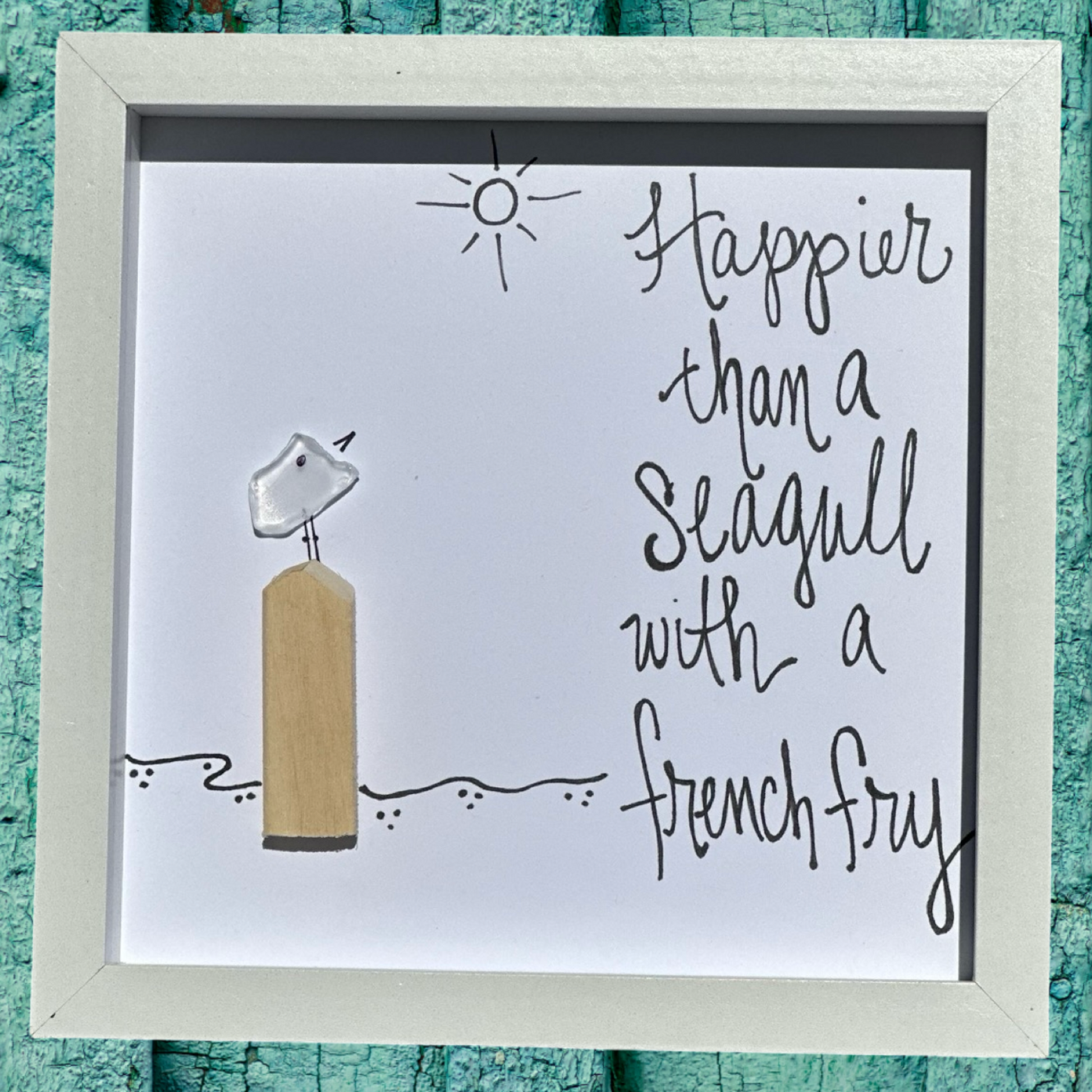 A framed shadowbox art piece with a seagull and a piece of seaglass, surrounded by a sun and ocean background, with the text 'Happier than a seagull with a French fry'.