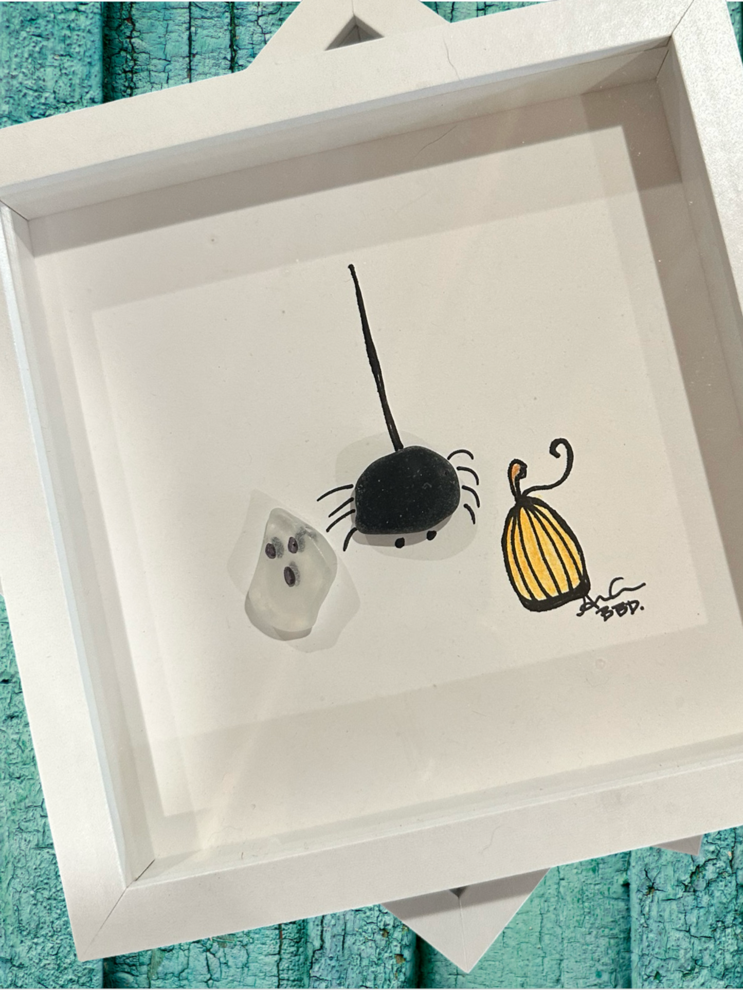 A decorative sea glass art piece featuring a drawing of a ghost, spider, and pumpkin, enclosed in a white frame.