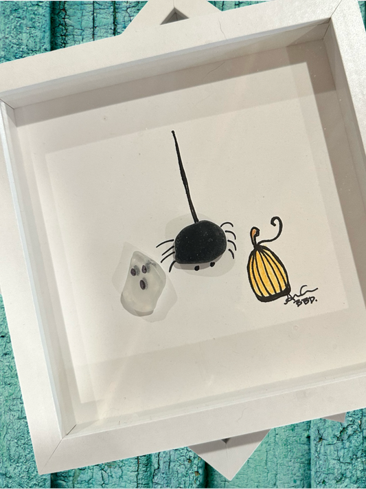 A decorative sea glass art piece featuring a drawing of a ghost, spider, and pumpkin, enclosed in a white frame.