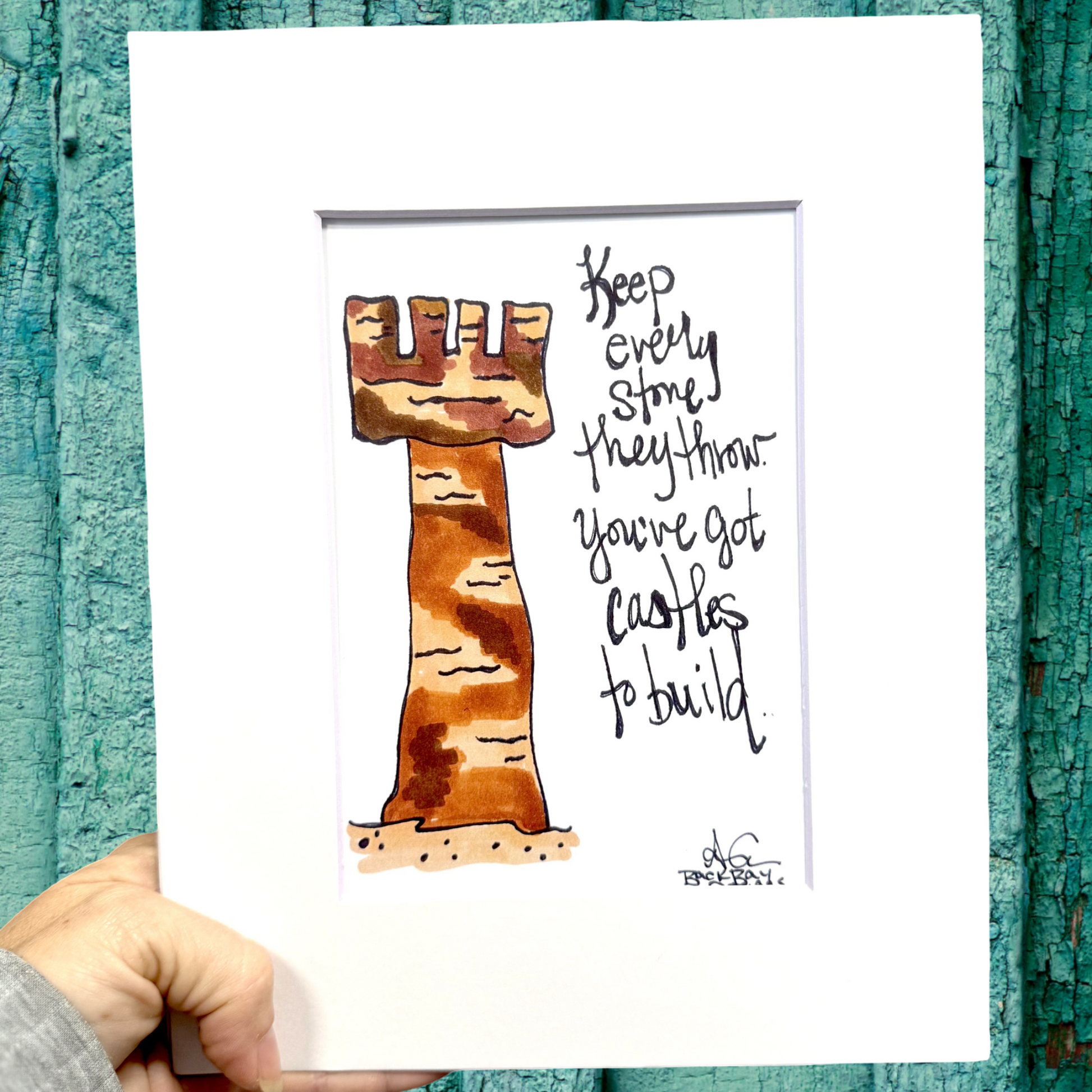 Hand holding a print with sand castle tower illustration and text against a textured teal background