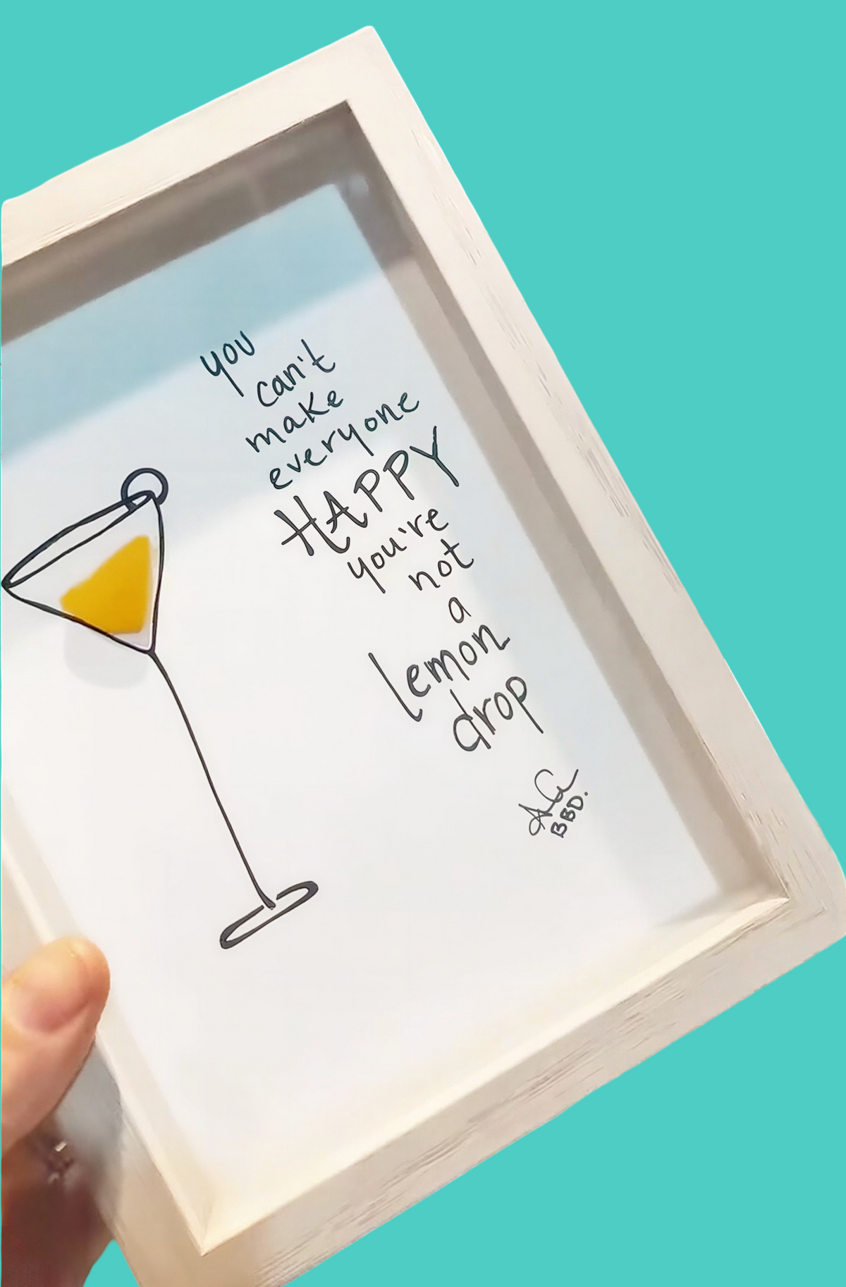 A hand holds a white-framed art print featuring a hand-drawn martini glass with a yellow liquid and the quote, 'you can't make everyone happy you're not a lemon drop' in black script on a turquoise background.
