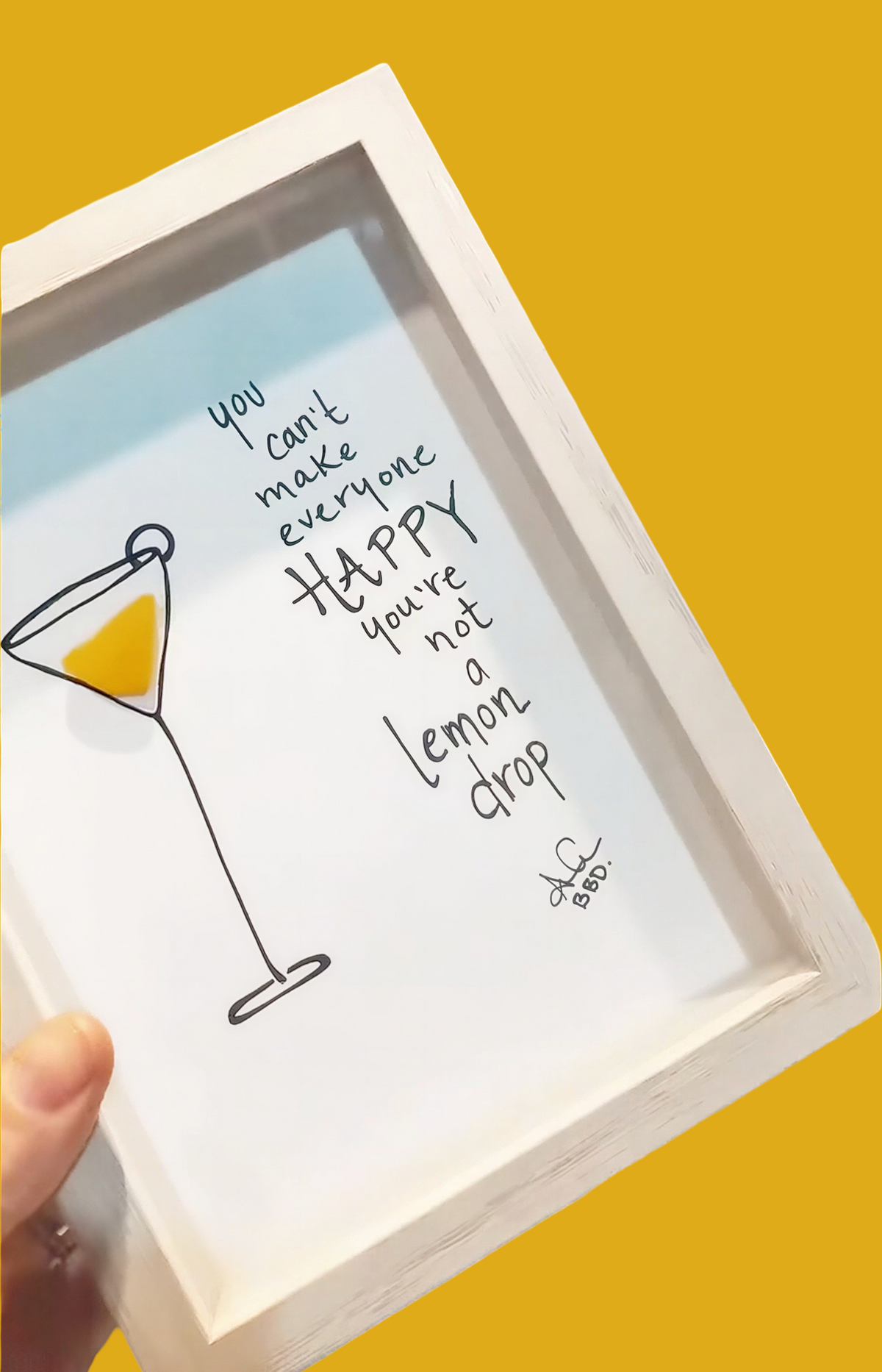 A hand holds a white-framed art print against a solid yellow background. The print features a hand-drawn illustration of a martini glass with a lemon slice and the quote, 'you can't make everyone happy you're not a lemon drop' in black script.