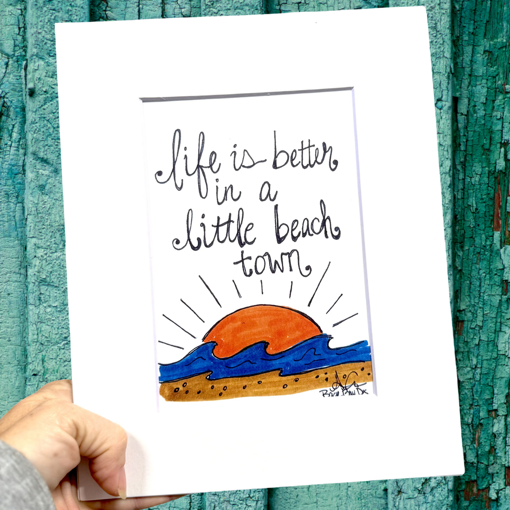 Hand holding a card with 'Life is better in a little beach town' text and beach graphic against a teal wooden background.