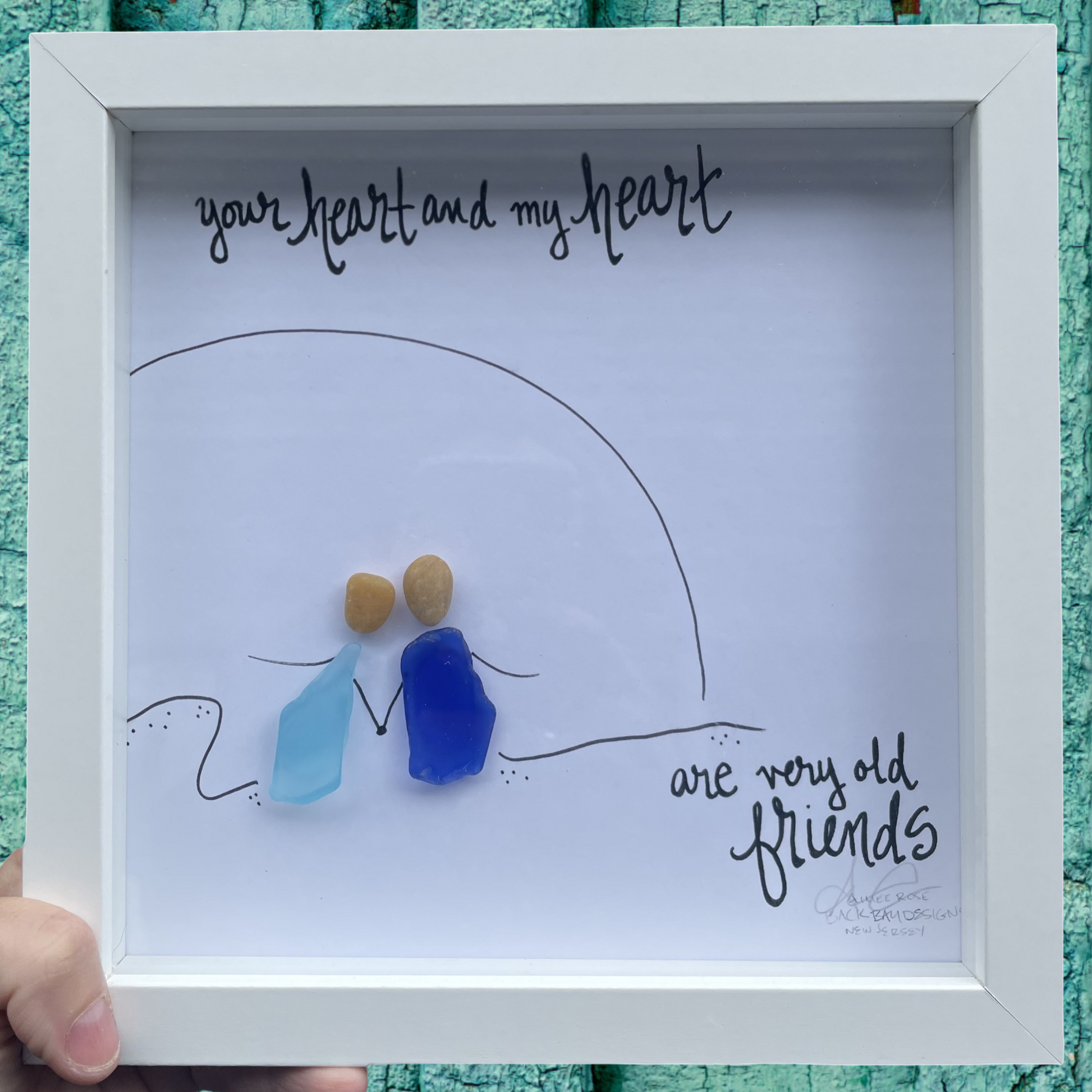 Framed artwork with two small figures made of blue and brown glass pieces, set against a white background with text. Your heart and my heart are very old friends. 