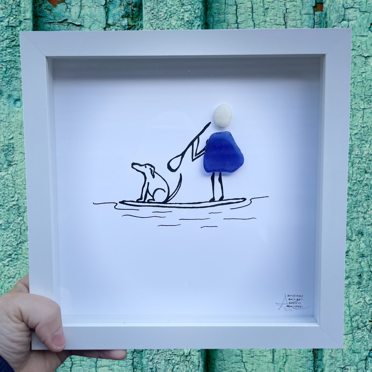 Framed artwork with a blue and black illustration of a person and dog on a surfboard against a teal textured background.