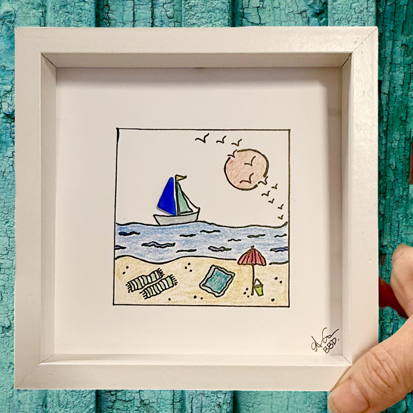 Framed sea glass art drawing of a beach scene with a sailboat, sun, and umbrella on a textured background. Sailboat Beach 6in MINI sea glass art on green