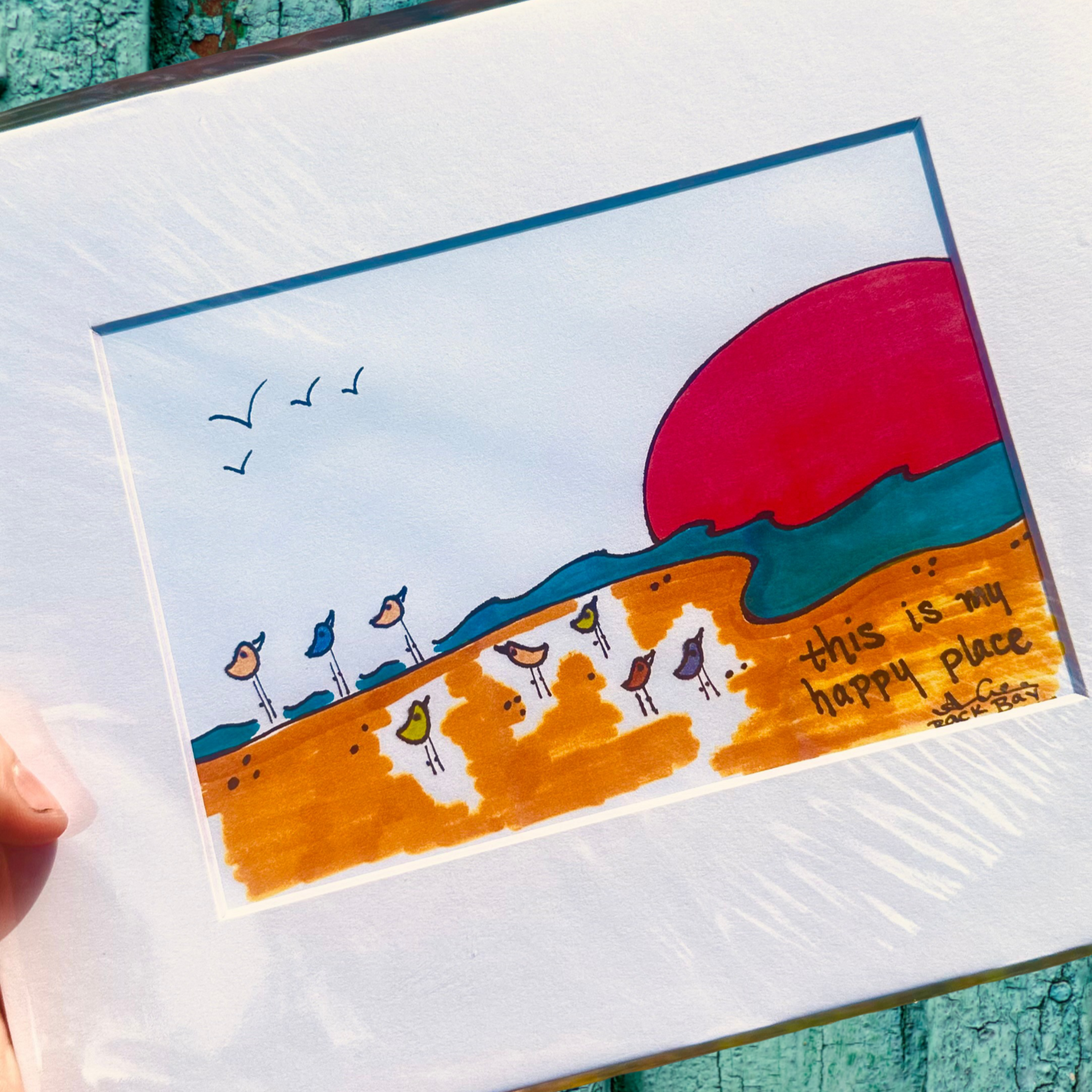 Sandpiper_Beach_Happy_Place_Print 
with a colorful illustration of birds and a sunset, held against a teal wooden background.