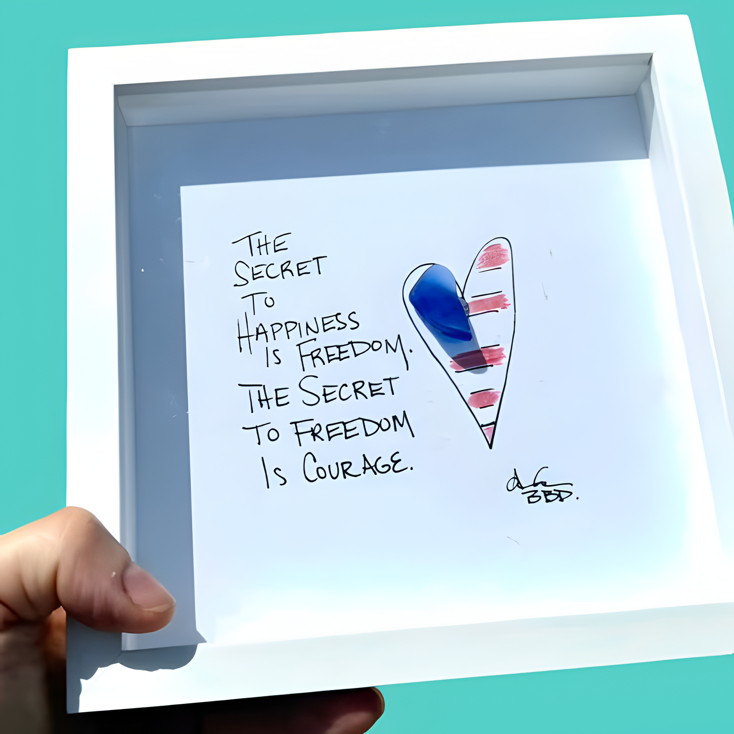 A hand holds a white square shadow box frame against a bright turquoise background. The frame contains a white card with the handwritten quote, 'THE SECRET TO HAPPINESS IS FREEDOM. The secret to Freedom is Courage." 