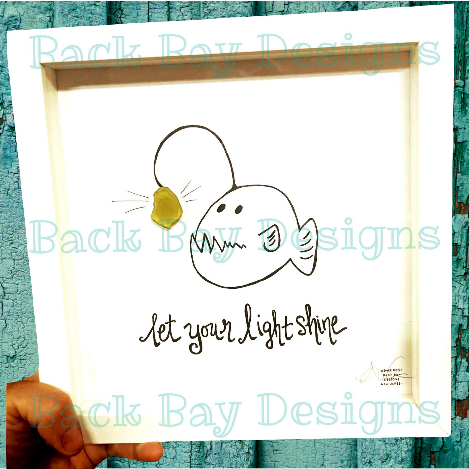 Framed artwork with a fish illustration and 'let your light shine' text, held against a textured teal background.