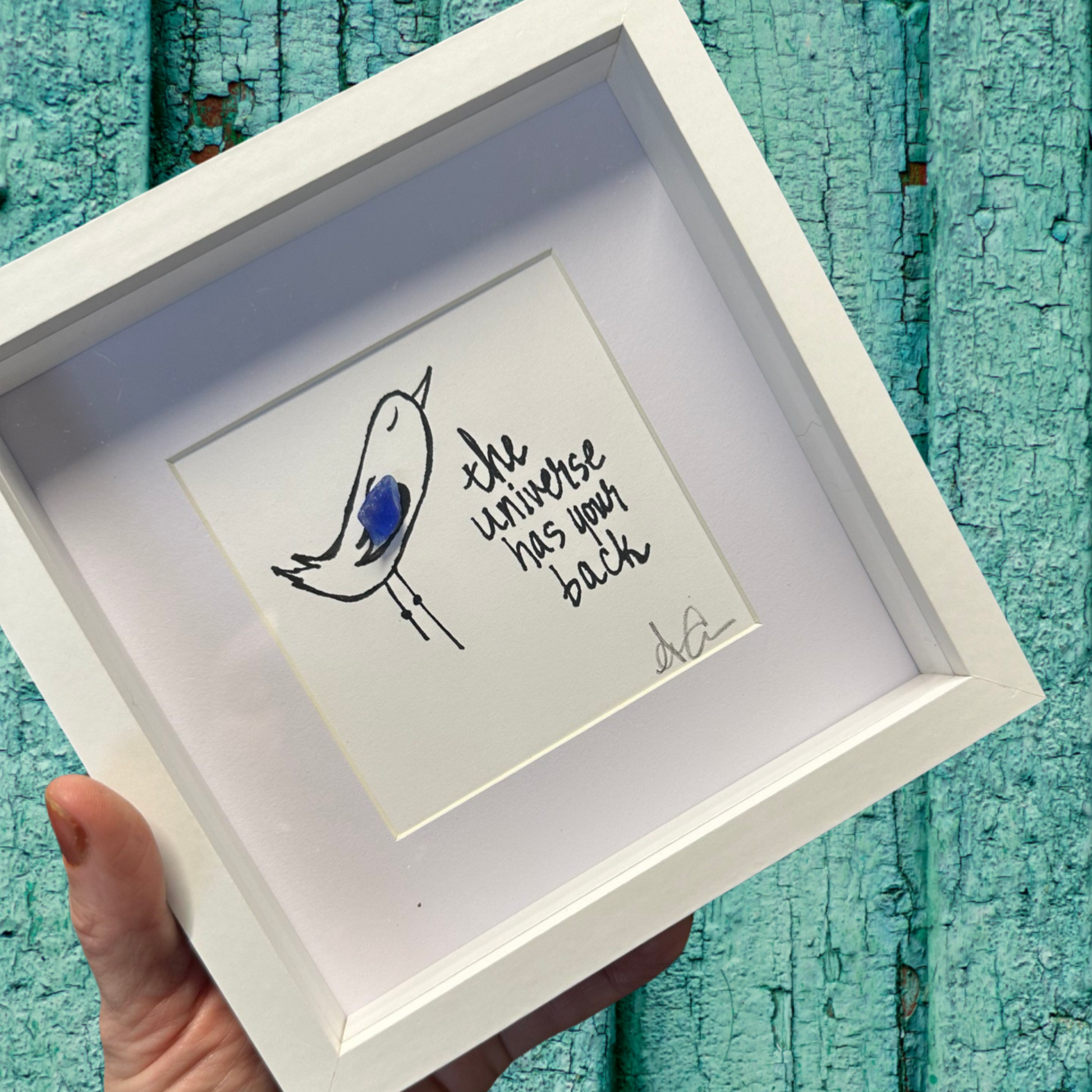 A 6x6 shadowbox with plexiglass, featuring a drawing of a bird and the text 'the universe has back'.
