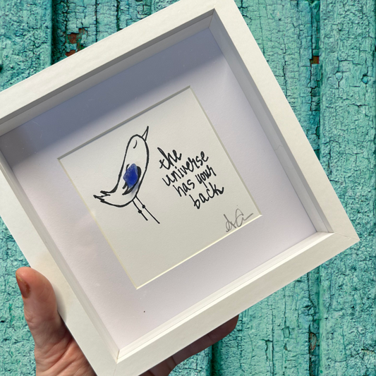A 6x6 shadowbox with plexiglass, featuring a drawing of a bird and the text 'the universe has back'.