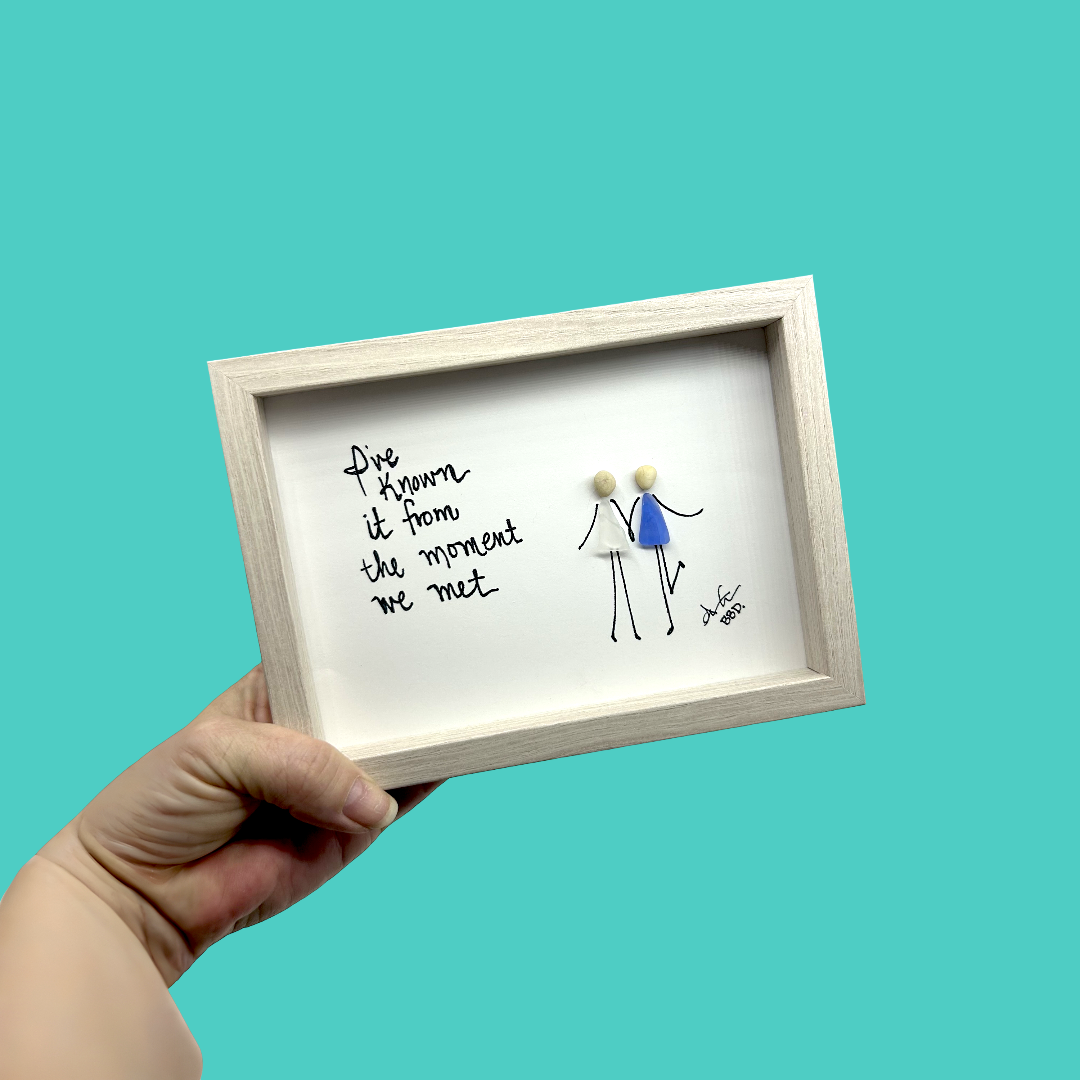 Quote: "I've Known it from the moment we met" Image: Two figures with sea glass dresses and pebble heads holding hands on hand-drawn stick arms and legs, one kicking up a leg