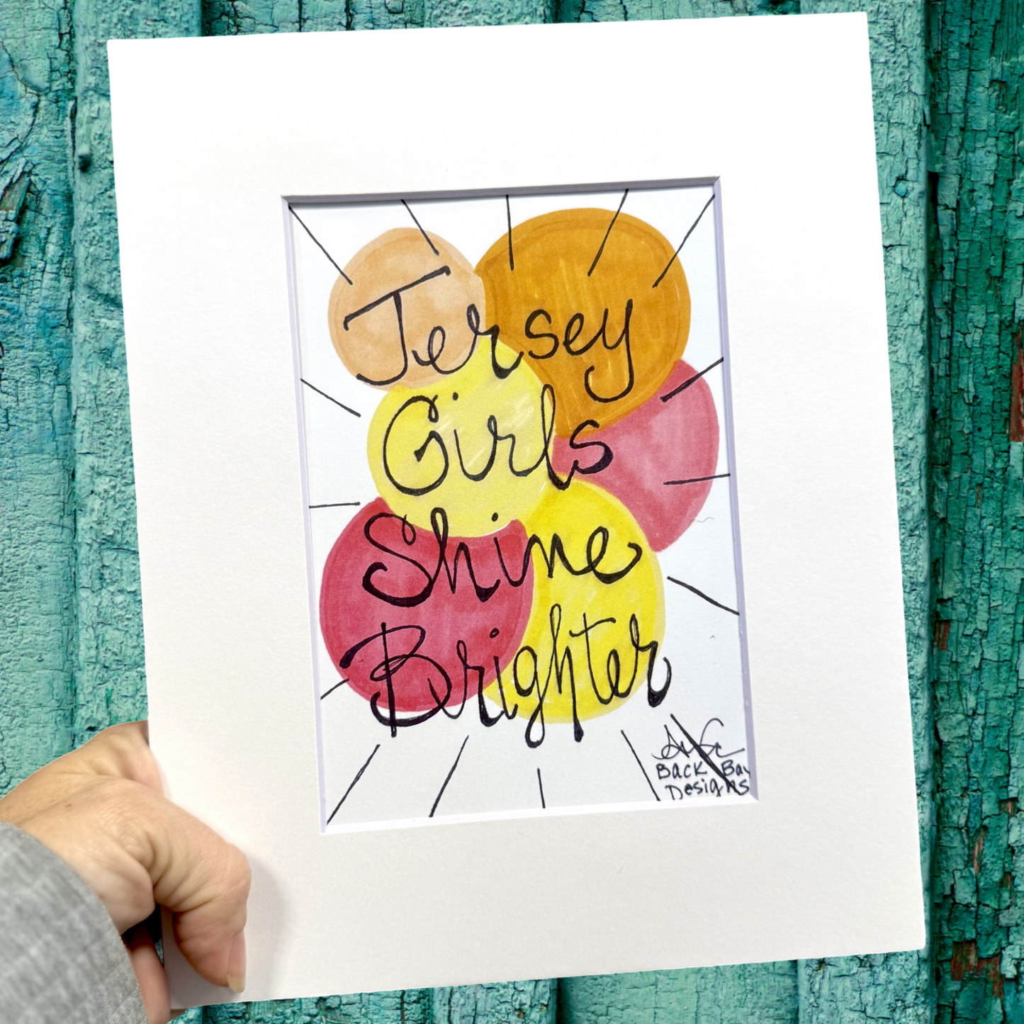 Hand holding a print with 'Jersey Girls Shine Brighter' text against a textured teal background