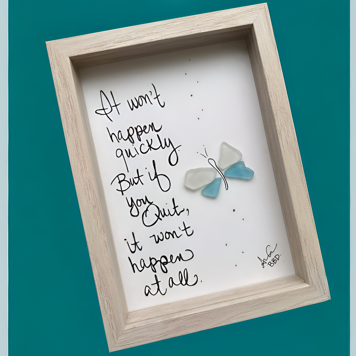 A square framed wall art print with a light wood frame, featuring a white background, black handwritten quote text, small black dots, and a small blue-and-white butterfly decoration. The frame is shown at an angle against a teal background.