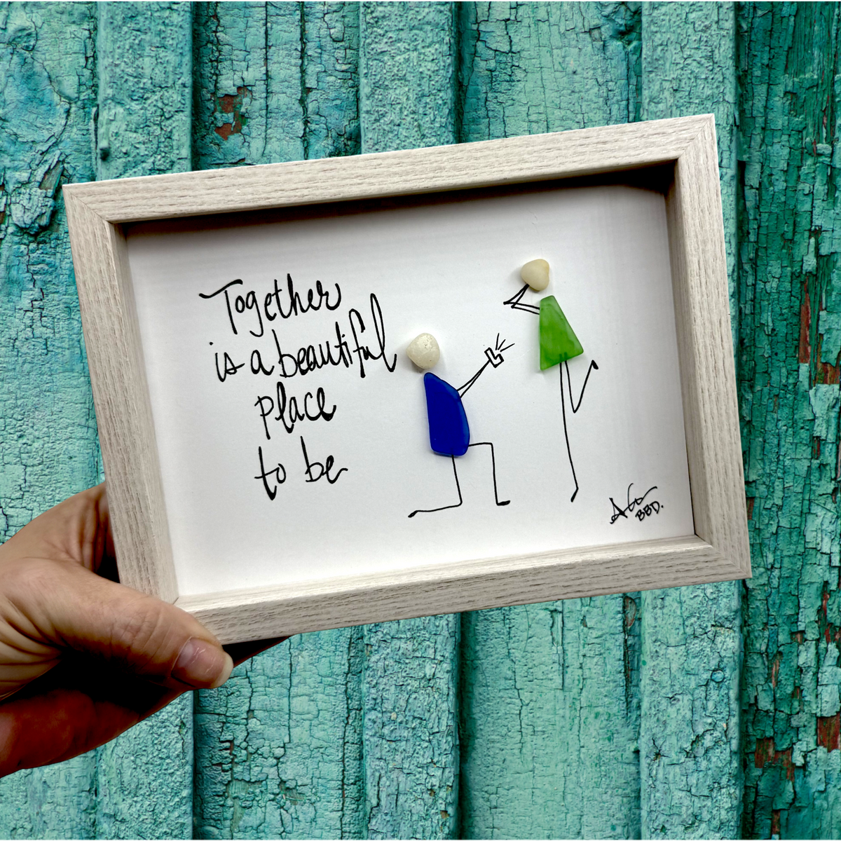 "Together is a beautiful place to be" Image: Sea glass couple with pebble heads and hand-drawn stick bodies, one on bended knee holding up a ring and the other with hand to face in surprise beach proposal