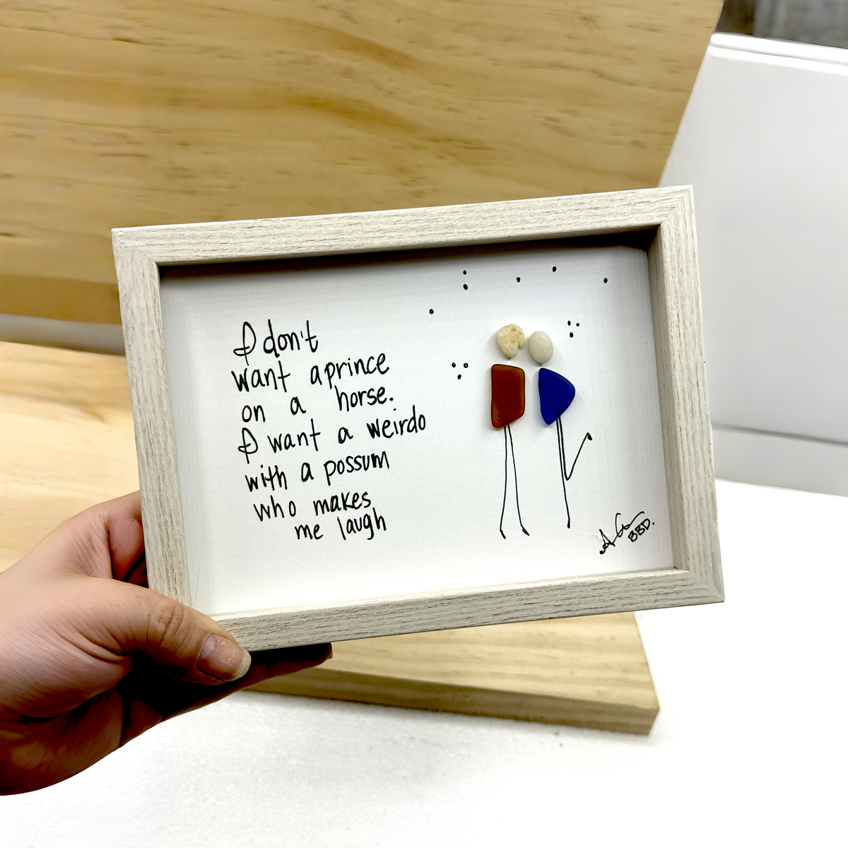 Quote: "I don't want a prince on a horse. I want a weirdo with a possum who makes me laugh" Image: Sea glass couple with pebble heads and hand-drawn stick bodies leaning in close, one kicking up a leg