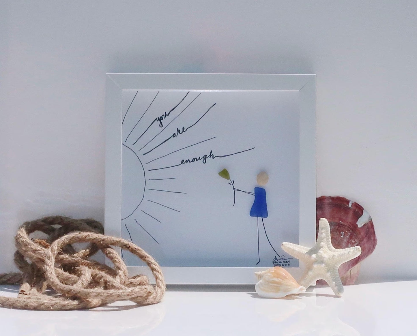 Framed artwork with motivational quote and stick figures, surrounded by natural elements on a light background.